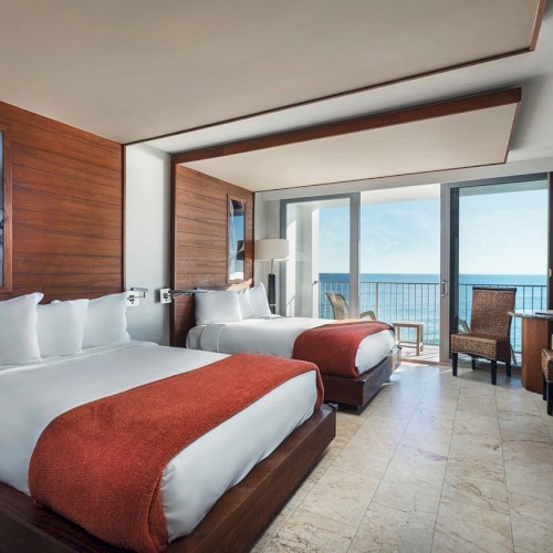 This image shows a modern hotel room with two double beds, a seating area, a flat-screen TV, and a balcony with a view of the ocean, ending the sentence.