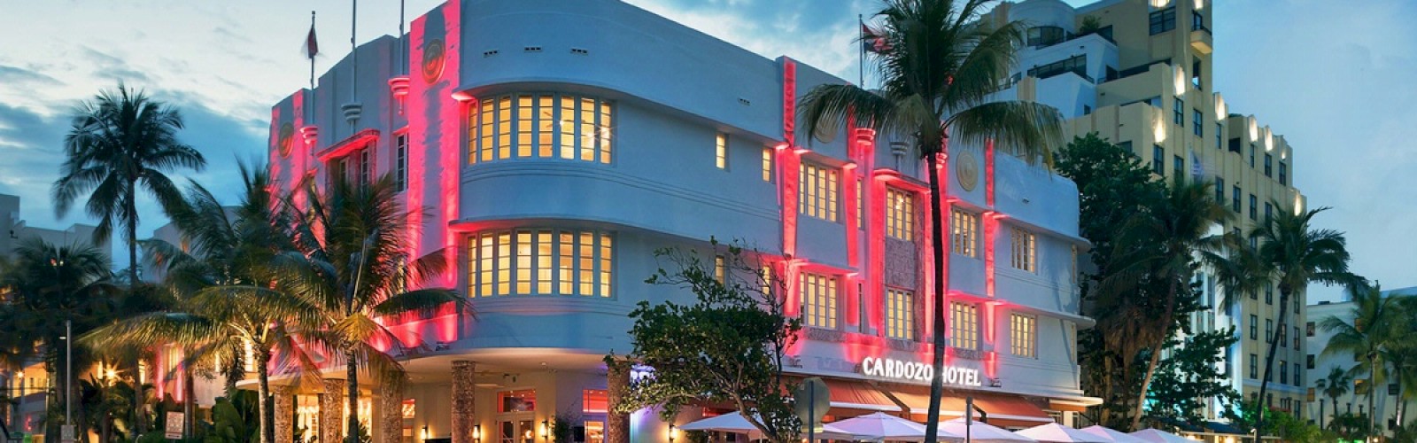 A hotel with vibrant red lights and a busy outdoor dining area on a tropical street lined with palm trees and parked cars.