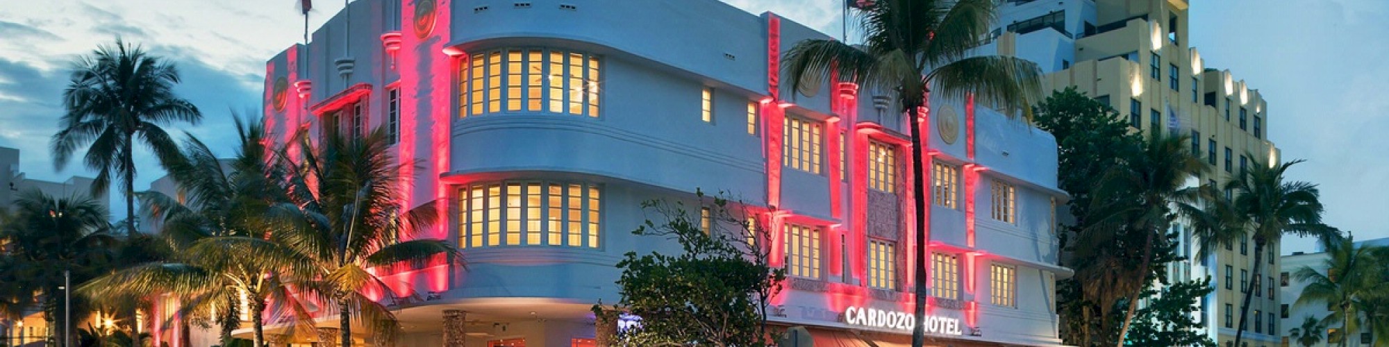 A hotel with vibrant red lights and a busy outdoor dining area on a tropical street lined with palm trees and parked cars.