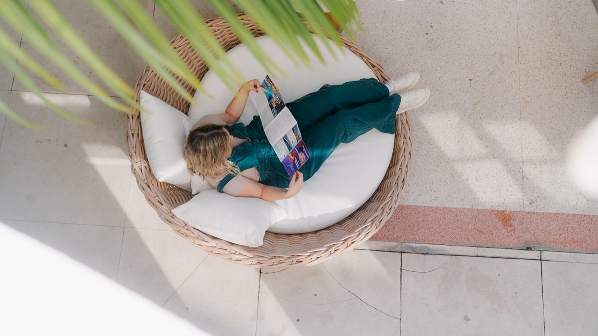 A person in a green outfit lounges in a round woven chair reading a magazine, with sunlight casting soft shadows on the tiled floor.