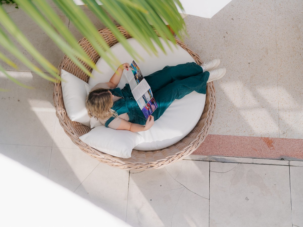 A person in a green outfit lounges in a round woven chair reading a magazine, with sunlight casting soft shadows on the tiled floor.