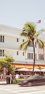 A white Art Deco building with American flags, palm trees, and a busy street scene in front.