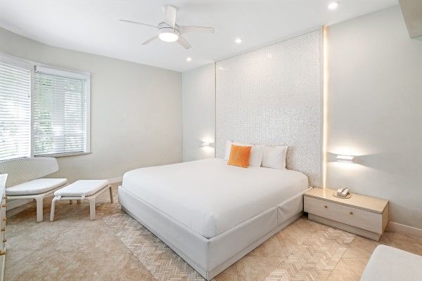 A modern bedroom with a large bed, white bedding, a single orange pillow, nightstands, a ceiling fan, and natural light through a window.