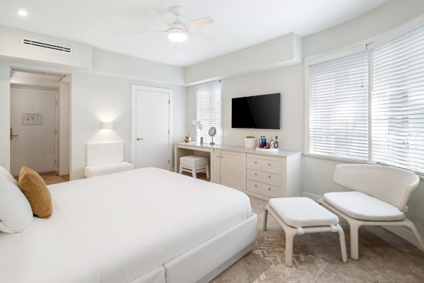 A modern bedroom with a white bed, a TV on a dresser, a chair with an ottoman, and large windows with blinds.