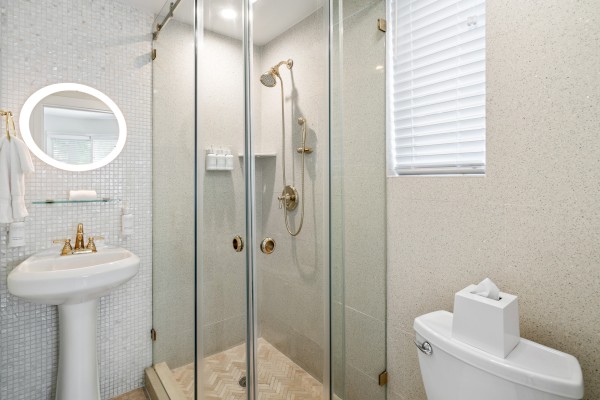 The image shows a modern bathroom with a glass shower, pedestal sink, and toilet, featuring gold fixtures and a round mirror.