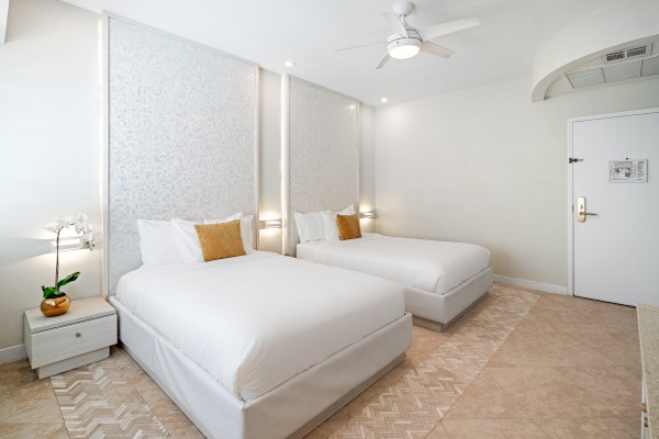 The image shows a modern hotel room with two white beds, a bedside table with a plant, and minimalistic decor, featuring a ceiling fan.