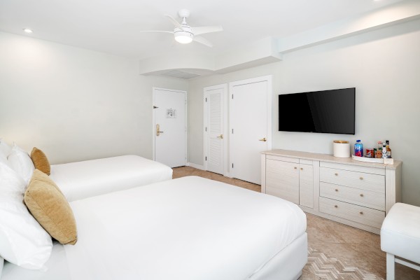 A hotel room with two beds, a TV on the wall, a dresser with snacks, and a door leading out. The decor is minimalist and modern.