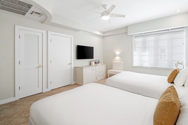 The image shows a clean bedroom with two beds, a TV on a dresser, two closed doors, and a window with blinds, creating a cozy atmosphere.