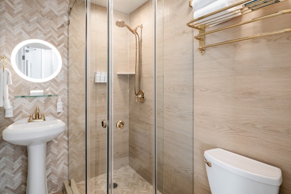 A modern bathroom with a glass shower, pedestal sink, toilet, and gold accents on fixtures and towel rack, featuring herringbone tiles.