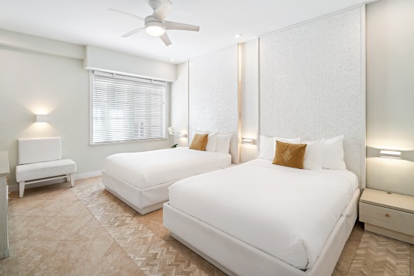 The image shows a modern bedroom with two double beds, white bedding, a ceiling fan, a window with blinds, and a neutral color scheme.