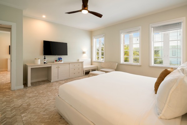 A bright, modern bedroom with large windows, a white bed, and minimal decor including a TV, ceiling fan, and cozy seating.