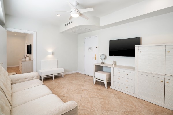 The image shows a modern, white living room with a sofa, TV, cabinet, and small desk with a mirror and stool.