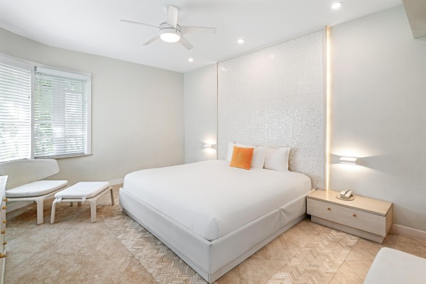 A bright, modern hotel-style bedroom with a large white bed, two nightstands, soft lighting, a ceiling fan, and a small seating area by the window, all in neutral tones.