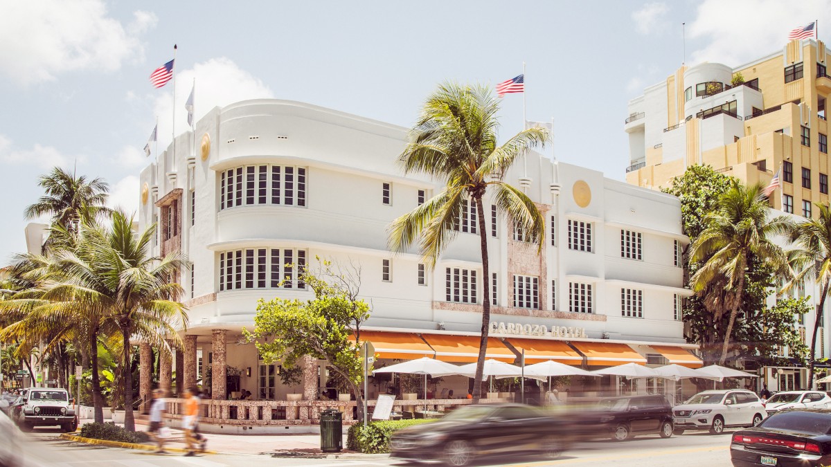 A white, multi-story art deco building stands on a busy street corner with palm trees and moving cars.