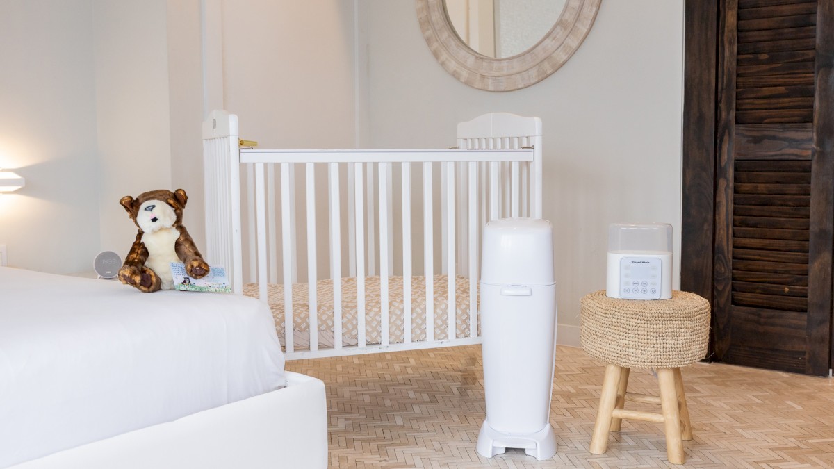 The image shows a cozy nursery with a white bed, a puppet resembling a bear, a white baby crib, a laundry bin, and a small woven stool.