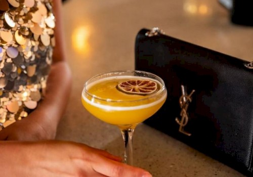 A person in a sequin dress holds a yellow cocktail with a dried citrus wheel, on a bar counter beside a black YSL clutch bag.