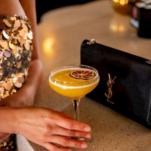 A person in a sequin dress holds a yellow cocktail with a dried citrus wheel, on a bar counter beside a black YSL clutch bag.