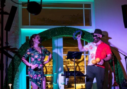A man and woman perform a comedy skit or act on a decorated stage with colorful lights, props, and microphones.