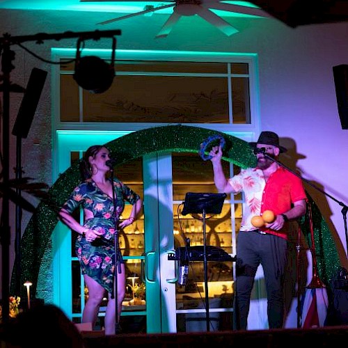 A man and woman perform a comedy skit or act on a decorated stage with colorful lights, props, and microphones.