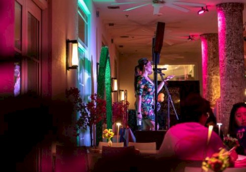 A woman performs stand-up comedy on stage in a dimly lit, colorful restaurant or cafe with vibrant neon lights and focused audience.