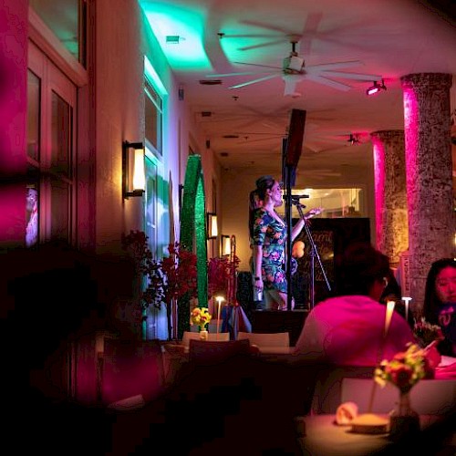 A woman performs stand-up comedy on stage in a dimly lit, colorful restaurant or cafe with vibrant neon lights and focused audience.