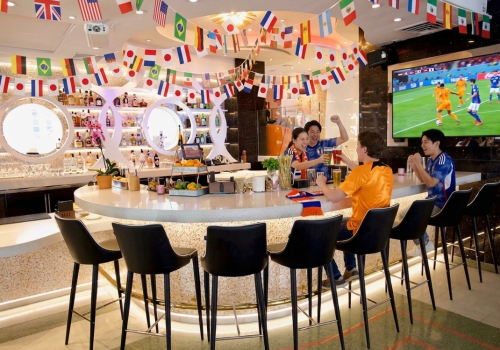 Friends cheer while watching a soccer game at a decorated bar with international flags, drinks, and a lively atmosphere.