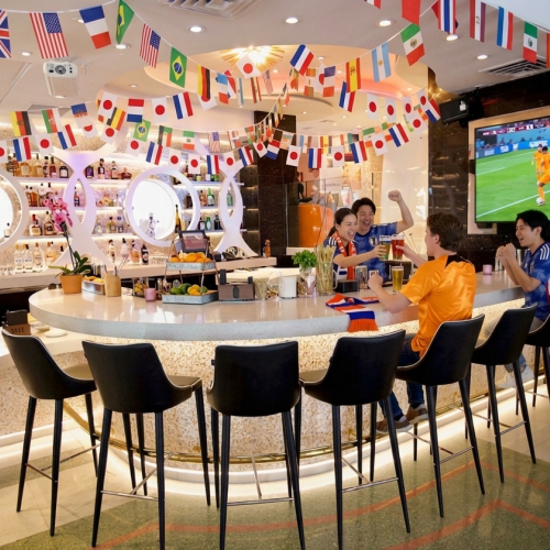 Friends cheer while watching a soccer game at a decorated bar with international flags, drinks, and a lively atmosphere.
