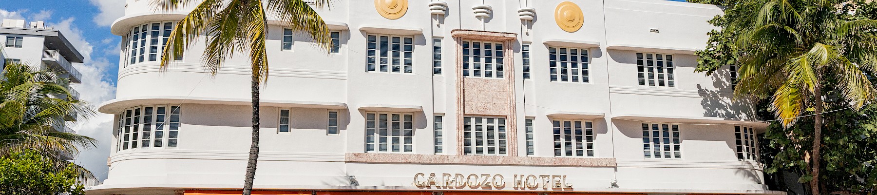 A white Art Deco building with orange awnings houses a cafe and outdoor seating, palm trees, and blue sky.