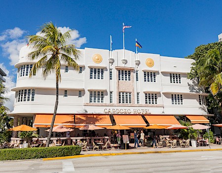 A white Art Deco building with orange awnings houses a cafe and outdoor seating, palm trees, and blue sky.